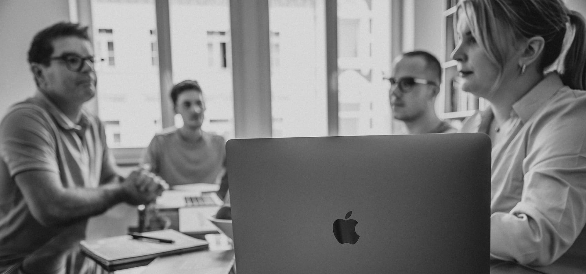 Ein Apple-PC steht im Vordergrund, während dahinter die Mitarbeiter am Tisch sitzen